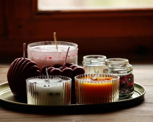 A variety of scented candles in glass jars on a tray by a window, creating a warm ambiance.
